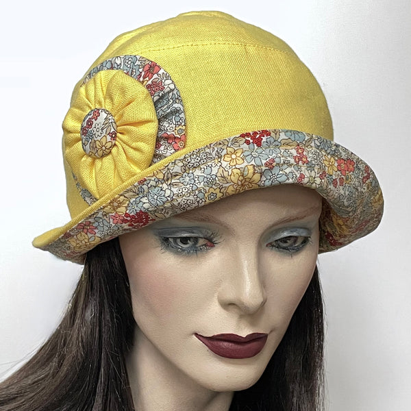 Find this handmade “Cloche” hat by local artisan Sue Scott at Eclection Ottawa. It is fashioned in mid-weight 100% linen in a lemon-yellow shade and the shape is a classic rounded cloche crown with a flexible brim that features a coordinated cotton floral in tones of chambray and soft corals on an off-white background at the under-brim. This hat is finished with hand-sewn double cockade trim in yellow linen and matching floral fabric and is fully lined with satin.  Size-medium: 22 ½.’’ 