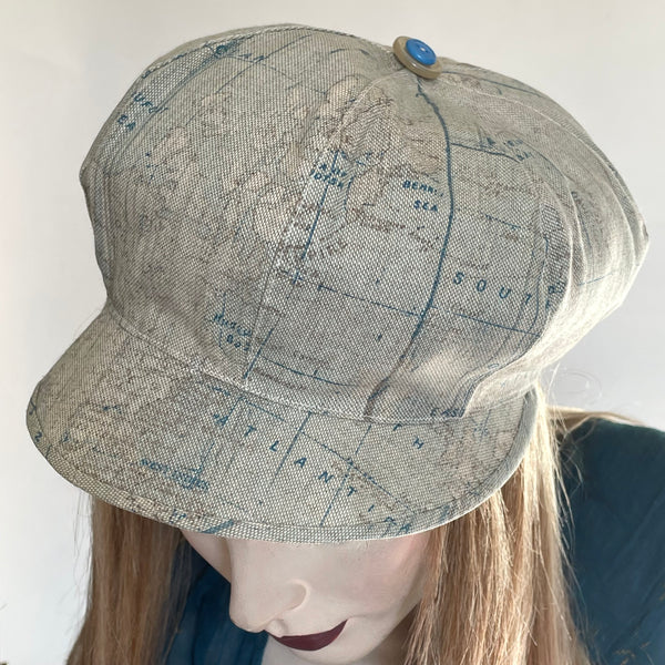 Find this handmade "Casquette" hat by local artisan Sue Scott at Eclection Ottawa. The fabric is a  mid-weight linen look cotton/polyester blend with a greige background enlivened by a decorative Map of the World printed motif in shades of taupe and blues. The shape is a classic eight-part voluminous crown, with a front peak. The trim is matching taupe and blue buttons on top. Fully lined. Size-medium: 22 1/2’’ with elastic in the back that gives it some give.