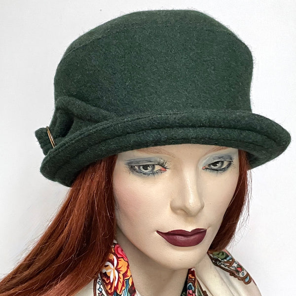 One-of-a-kind, handmade “Cloche” hat by Ottawa artisan Sue Scott. The cloche is fashioned in cozy forest green wool and has a handmade bow trim finished with topstitching and a vintage buckle at its center. This hat embodies the beautiful 1920s classic style and adds a modern twist.  It is lined with a satiny, flannel-backed Kasha lining that’s easy on the hair, stops wind for added warmth, and features a flexible brim that can be styled as you like. Size-medium: Approximately 22 ½.’’ Dry Clean