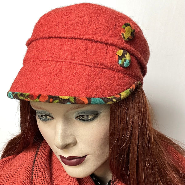 This one-of-a-kind hat, handmade by local artisan Sue Scott, is called the "Crumper".  This cap is fashioned in a pumpkin wool blend fabric with a rich boucle texture. The shape is a straight-sided, flat-topped crown, with a front peak. It is embellished with  button trims covered in velveteen print in shades of teal, green and orange on a brown background. It features the same print fabirc at the underbrim.  Fully lined with windproof lining. Size-medium, 22 1/2" with an elastic at the back.