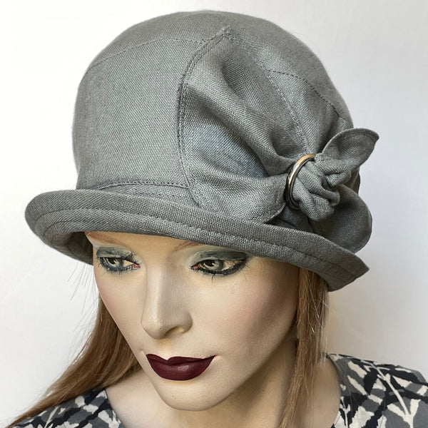 Find this Cloche hat at Eclection Ottawa. It's a charming shape that embodies the beautiful 1920s classic style and adds a modern twist to it. It is fashioned in medium-weight 100% linen in a classic medium grey shade and the shape is a classic rounded cloche crown that features a flexible top-stitched brim that you can wear as you please.  This hat is finished off with a large matching hand-sewn bow trim with a vintage buckle at its center and is fully lined.  Size-medium: approximately 22 ½.’’