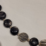 Closeup of strand of flat round black hyperstein beads with shiny finish spaced with small silver beads with swirl texture and the featured larger silver beads with swirl pattern 