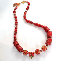 This one-of-a-kind necklace is handmade by Ottawa artisan Khalia Scott. It is carefully crafted out of tinted coral, large and beautifully textured lampwork beads, small glitter glass beads in a rich tone of red, tinted agate and finished off with a gold plated bee charm, faceted separator beads and findings. It is strung securely by hand on a heavy coated multi-strand tiger tail cord and has a gold-plated clasp. It is measuring 25 3/4".