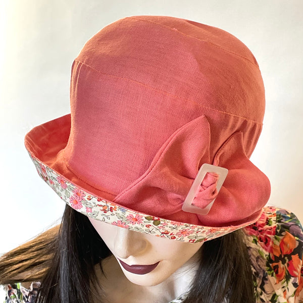 Find this handmade “Cloche” hat by local artisan Sue Scott at Eclection Ottawa. It is fashioned in medium-weight linen in a soft cameo pink shade. The shape is a classic rounded cloche crown that features a flexible brim that can style to your tastes. It is finished off with large bow trim and a vintage buckle at its center and cotton floral fabric in matching colour at the under brim. Fully lined. Size medium, approximately 22 ½.’’ 
