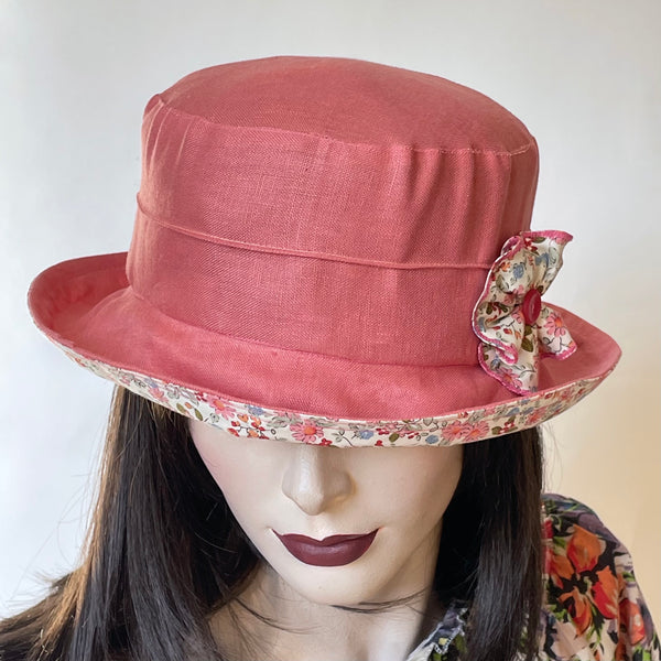 Find this handmade "Jojo" hat by local artisan Sue Scott at Eclection Ottawa. The fabric is a mid-weight 100% linen in a soft cameo pink shade that brings a pop of colour in the warmer season. The shape is a straight-sided crown with a flat top and a medium brim that curves upwards and can be positioned and show the cotton floral of the under brim. It is finished off with a rosette in the same floral cotton and a matching button at its center. Fully lined. Size-medium: approximately 22 1/2’’