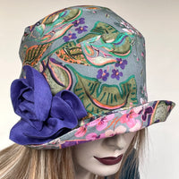 Find this handmade “Cloche” hat by local artisan Sue Scott at Eclection Ottawa. It is made in a rich mid-weight 100% linen fabric with a grey background enlivened by a decorative tropical floral motif in shades of pinks, greens, purples and oranges. The shape is a rounded cloche crown that features a brim that can be styled to your tastes.  This hat has a voluminous, hand-sewn flower trim in matching purple fabric as a finishing touch and is fully lined.  Size-medium: approximately 22 ½.’’