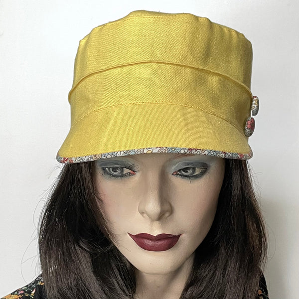 Find this Crumper cap handmade by artisan Sue Scott at Eclection Ottawa. It is fashioned in mid-weight 100% linen in a beautiful lemon-yellow shade. The shape is a straight-sided, flat-topped crown, with a classic front peak that features a floral under-peak in coordinated cotton fabric in tones of chambray, yellow and soft corals on an off-white background. It is finished off with a covered button trim that repeats the fabric of the under-peak. Fully lined. Size-medium 22 1/4’’