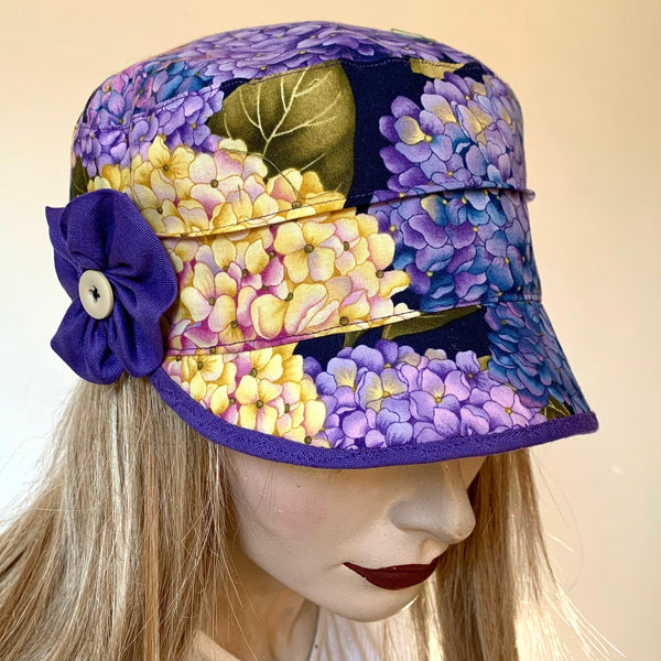 Fanfreluche Crumper Hat in Hydrangea Cotton Print in Purples, Yellow, pink and Green Colours, with a Purple Rosette Trim. The shape is a straight-sided, flat topped crown, with a classic front peak. Original and handmade in Canada Hat