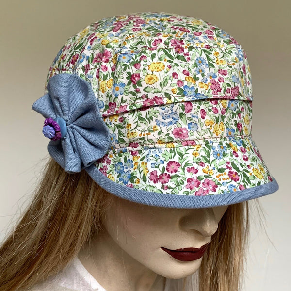 Fanfreluche Crumper Hat in Garden Floral  Cotton Print  of Blues, Pinks and Greens with Blue Rosette Trim.The shape is a straight-sided, flat topped crown, with a classic front peak . Original and handmade in Canada hat.