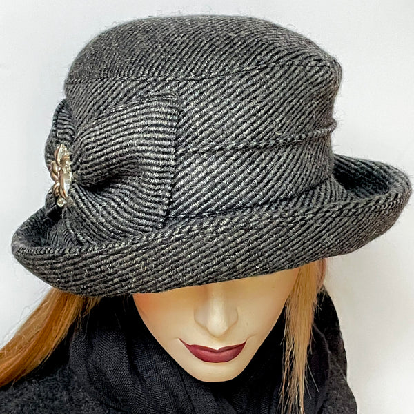 Find this one-of-a-kind, handmade “Jojo” hat with a Stella brim by Ottawa artisan Sue Scott at Eclection Ottawa. It is fashioned in a grey and black striped wool and cashmere blend fabric that adds texture to this classic hat. The shape is a straight-sided crown with a flat top and a Stella brim that is flexible and can be styled easily. Hand-sewn flat bow on the side with a vintage metal ornament at its centre. Windproof Kasha lining for added warmth. Size-medium: 22 ½.’’