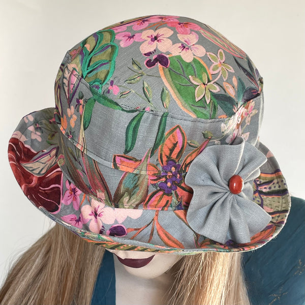 Find this handmade "Jojo" hat by local artisan Sue Scott at Eclection Ottawa. The fabric is a mid-weight 100% linen fabric with a grey background enlivened by a decorative tropical floral motif in shades of pinks, greens, purples and oranges. The shape is a straight-sided crown with a flat top and a medium brim that curves up and can be positioned. Finished off with a hand-sewn rosette in matching grey fabric with a vintage button at its center. Fully lined. Size-med: approximately 22 1/2’’