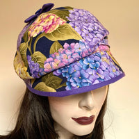 Fanfreluche Miss Kitty Hat in Hydrangea Cotton Print in Purples, Yellow, Pink and Green colours, with a Peak and a Purple Bow on Top. Original and handmade in Canada hat.