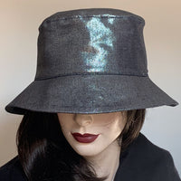 Find this handmade bucket hat by local artisan Sue Scott at Eclection Ottawa. The "Nina" is made off a cotton/poly silver denim fabric with a hint of lurex for a shiny metallic look. The shape is a classic bucket with a straight-sided crown and a flat top, with a medium brim that angles down  or can be  curved up. Fully lined. Size 22 1/2’’