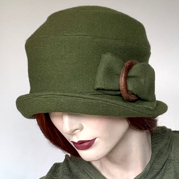 One-of-a-kind, handmade “Cloche” hat by Ottawa artisan Sue Scott. It is fashioned in a cozy wool / polyester blend boiled wool in a lovely olive colour. It is beautifully finished off with a wooden vintage buckle trim. It embodies the beautiful 1920’s classic style and adds a modern twist to it. It is lined with satin and features a flexible brim that you can style to your tastes. Size-medium: 22 ½.’’ 