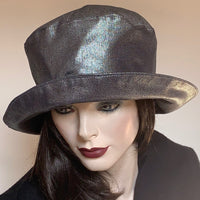 This hat, handmade by local artisan Sue Scott, is called the "Over the Topper". This hat is fashioned in a cotton/polyester silver denim fabric with a hint of lurex for a shiny metallic look that mimics the reflections on the surface of a lake. The shape has volume with a crown that flares at the top and a wide brim that curves upwards and can be positioned. Fully lined with red satin. Size-medium/large: 22 3/4’’