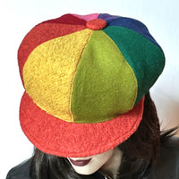 This Casquette is fashioned in a mix of different fabrics that goes from wool flannel,  cashmere and wool blend to 100% wool coating. The shape is an eight-part crown, with some volume and a front peak where each part of the crown is made with a different colour. Red, candy pink, purple, forest green, indigo blue, chartreuse, yellow and Orange. Orange peak and covered button on top. Wind-resistant lining. Size M/L: 22 1/2’’ with an elastic in the back. 
