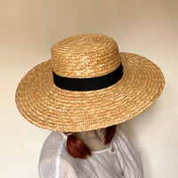 Find this flattering and elegant boater hat at Eclection Ottawa. The “Florida” is crafted out 100% braided straw and trimmed with a black polyester grosgrain ribbon and a flat bow on the side. The shape is an oval flat top crown with straight sides and a flat, straight brim. Size is 22 1/2" adjustable with an elastic inside the grosgrain interior  band. Brim 4 1/4". Crown 3 1/2"