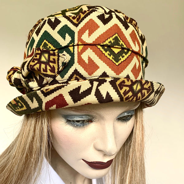 This is a classic wearable style with a funky twist, handmade “Jojo” hat by Ottawa artisan Sue Scott. Fashioned in a cotton/polyester print with a cream background that boasts a fun tribal print in tones of buttery yellow, rust, plum and greens. It is finished off with a handmade bow trim in matching fabric. It has a flexible brim that can be styled easily. Fully lined with satin. Size-medium: 22 ½.’’