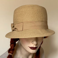 Tan tweed colour "Millie" Cloche Hat made of paper braid with 4 1/2" deep rounded crown.  Downward Brim of 2" in front and 3/4" in back. Tan colour Grosgrain ribbon trim. Interior band with adjustable elastic. UPF 50+ and size medium- 22" at Eclection Ottawa