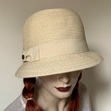 White tweed colour "Millie" Cloche Hat made of paper braid with 4 1/2" deep rounded crown.  Downward Brim of 2" in front and 3/4" in back. Sand colour Grosgrain ribbon trim. Interior band with adjustable elastic. UPF 50+ and size medium 22".  at Eclection Ottawa