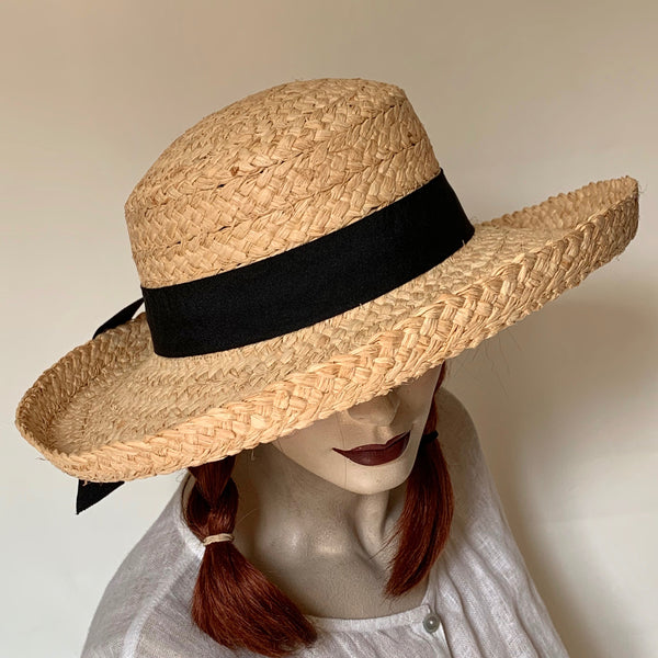 Find this flattering and charming hat at Eclection Ottawa. The Rachel is crafted out of sewn 100% braided natural raffia and is finished off with a large grosgrain ribbon trim tied in a knotted bow with tails at the back. The shape is an oval cloche crown of approximately 4" deep, and a  kettle brim of around 3" wide with a 1" edge that flips up. The interior band is made of a soft large elastic for comfort and adjustability. Size medium at approximately 22 1/2".