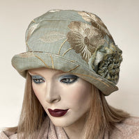 Find this handmade “Cloche” hat by local artisan Sue Scott at Eclection Ottawa. It is fashioned in a rich mid-weight cotton/polyester blend tapestry fabric with a seafoam and champagne background that is enlivened by a floral motif in shades of beige, light taupe and champagne. The shape is a classic rounded cloche crown that features a top-stitched brim that can be styled to your tastes.  This hat has a large, hand-sewn flower trim and is fully lined.  Size-medium: approximately 22 ½.’’ 