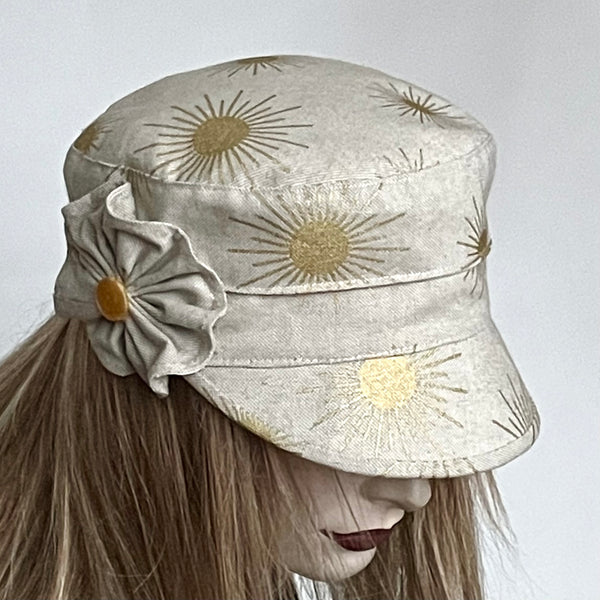 This hat, handmade by local artisan Sue Scott, is called the "Crumper". The fabric is a  mid-weight linen look cotton/polyester blend with a natural background and is enlivened by decorative gold suns printed motif. The shape is a straight-sided, flat-topped crown, with a classic front peak (that sports the same fabric on the underside) and it is embellished with a hand-sewn rosette with a button at its centre. Fully lined with dupioni silk. Size-medium 22 1/4’’