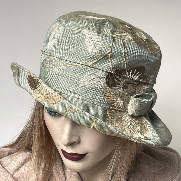 Find this handmade "Jojo" hat by local artisan Sue Scott at Eclection Ottawa. The fabric is a rich mid-weight cotton/poly blend tapestry fabric with a seafoam and champagne background that is enlivened by decorative floral motifs in shades of beige, light taupe and champagne. The shape is a straight-sided crown with a flat top, with a medium brim that curves upwards and can be positioned. It is finished off with a hand-sewn tailored bow in the same fabric and is fully lined. Size-med: 22 1/2’’