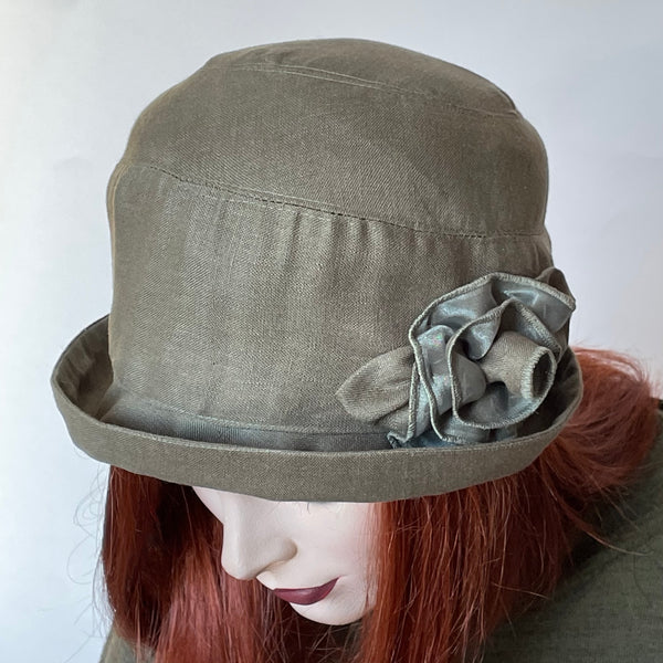 One-of-a-kind, handmade “Cloche” hat by Ottawa artisan Sue Scott. It's a charming shape that provides sun protection with style and embodies the beautiful 1920’s classic style with a modern twist. It is fashioned in lightweight linen in easy to wear sage green shade. The shape is a classic rounded cloche crown that features a flexible brim that can style to your tastes.  This hat is finished off with a voluminous matching hand-sewn rose trim and is fully lined with satin.  Size-medium: 22 ½.’’
