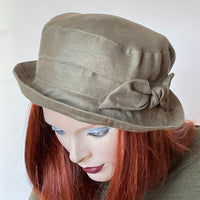 This one-of-a-kind hat, handmade by local artisan Sue Scott, is called the "Jojo". It's a practical and elegant shape that provides sun protection with style. The fabric is 100% lightweight linen in a versatile and easy-to-wear sage green shade. The shape is a straight-sided crown with a flat top with a medium top-stitched brim that curves upwards and can be positioned. It is finished off with a hand-sewn bow and is fully lined with Bemberg lining. Size-medium: 22 1/2’’