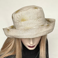 Find this "Thomasina" handmade sun hat by local artisan Sue Scott at Eclection Ottawa. It's a practical and elegant shape that provides great sun protection with style.  The fabric is a mid-weight linen look cotton/polyester blend with a natural background that is enlivened by decorative gold suns printed motif. The shape is a straight-sided 3 part crown with a flat top and a generous 3 part kettle brim of 2 1/2" wide with a 2" edge that flips up. Fully lined with rayon. Size-medium: 22 1/2’’